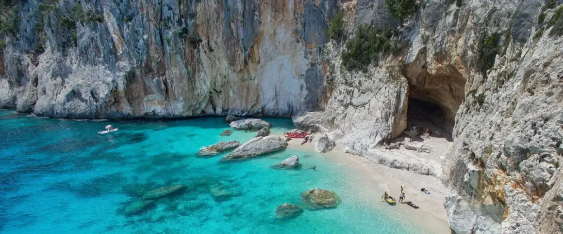 Spiaggia incantevole sulla costa di Baunei, con acque cristalline, rocce imponenti e una grotta marina. Kayak e persone godono del sole.