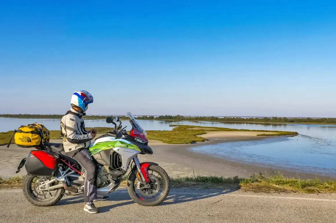 Motociclista con moto da enduro ferma sul ciglio di una strada sterrata, con sullo sfondo il paesaggio del Delta del Po.