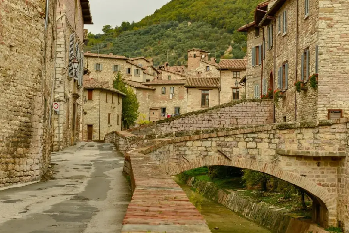 Antico ponte in pietra attraversa un ruscello, fiancheggiato da case in pietra e vicoli stretti, tipici dei paesi in Umbria.