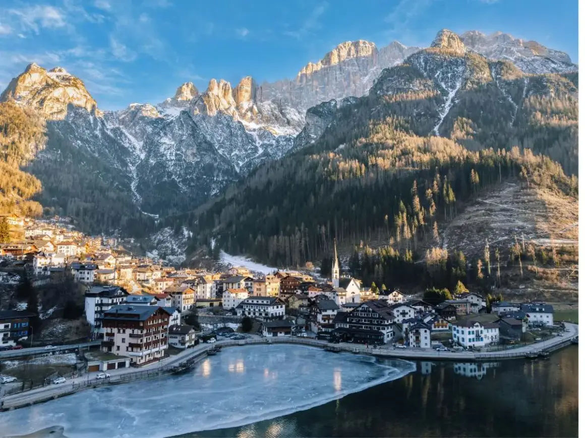 Villaggio alpino innevato ai piedi delle Dolomiti, ideale per escursioni invernali. Lago ghiacciato riflette le montagne.