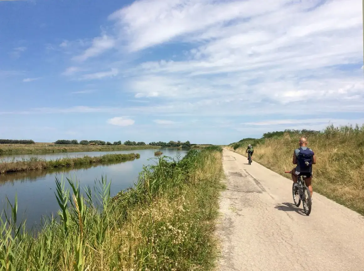 Cicloturismo in Emilia Romagna: due ciclisti pedalano su una strada sterrata lungo un canale, con vegetazione rigogliosa e cielo azzurro.