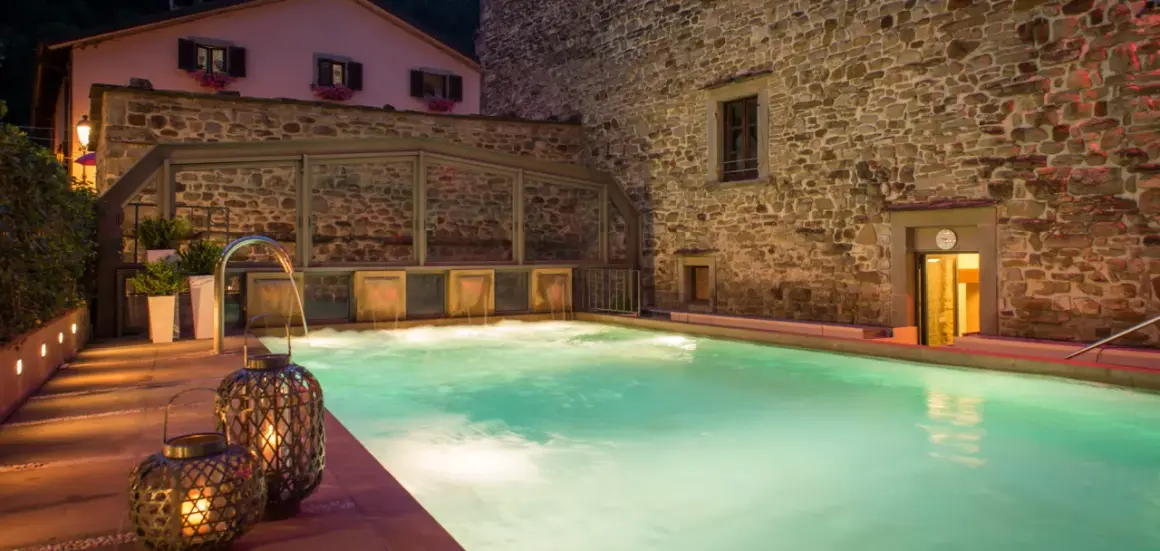 Piscina termale illuminata da lanterne, con cascate d'acqua e un edificio in pietra. Un'oasi di relax che ricorda le terme della Riviera Romagnola.