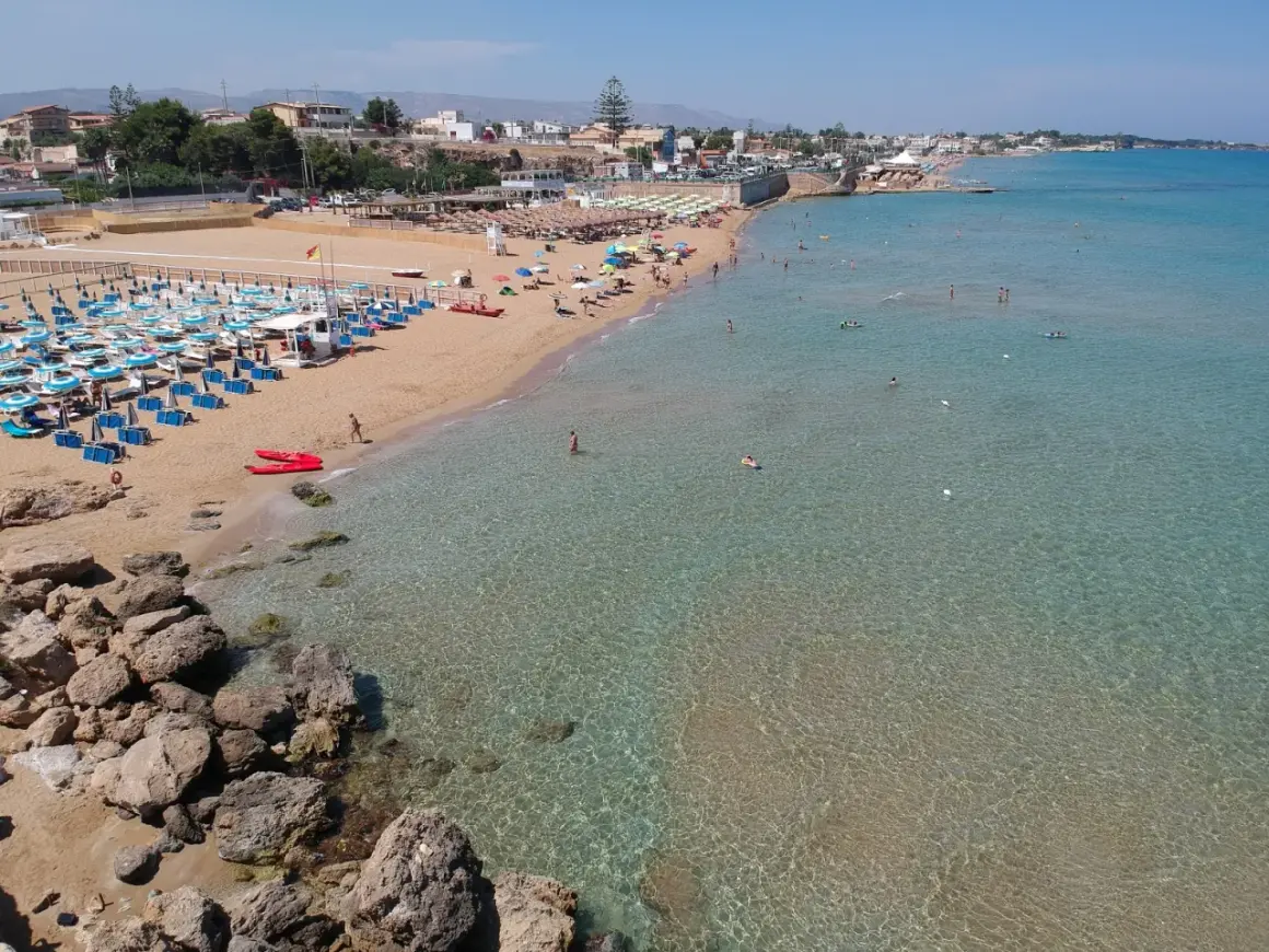Una spiaggia affollata con lettini e ombrelloni, bagnanti in acqua e due kayak rossi sulla sabbia. Il noto mare cristallino invita a un tuffo.