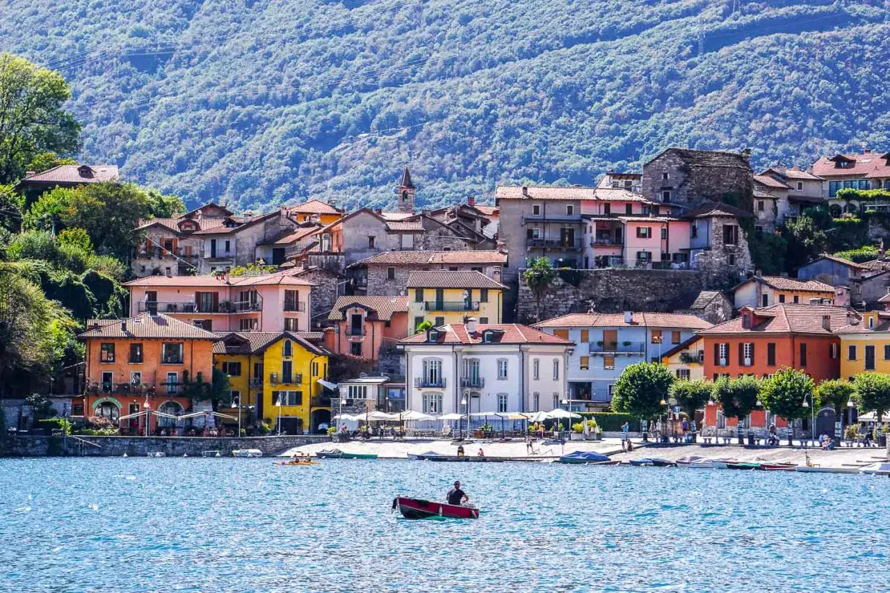 Un uomo in barca sul lago, con case colorate che si arrampicano sulla collina. Questo è un scorcio di dove si trova Domodossola, un luogo incantevole.