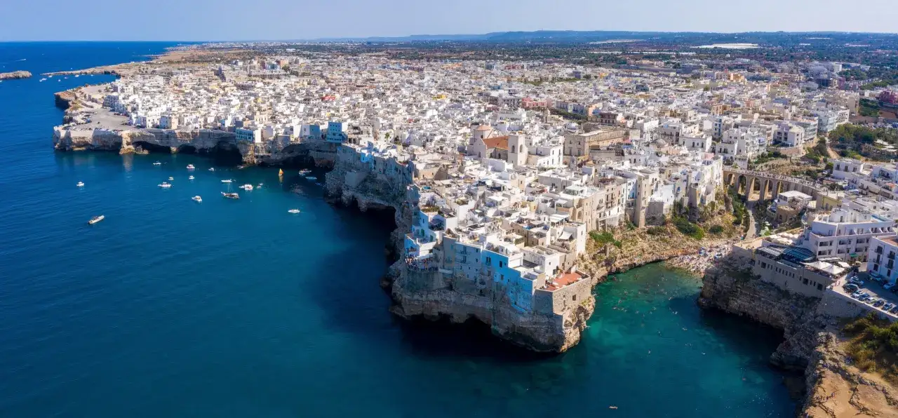 Panorami mozzafiato della Valle d'Itria con il mare blu intenso che lambisce scogliere bianche. Case a picco sul mare, barche e gente che si gode il sole.