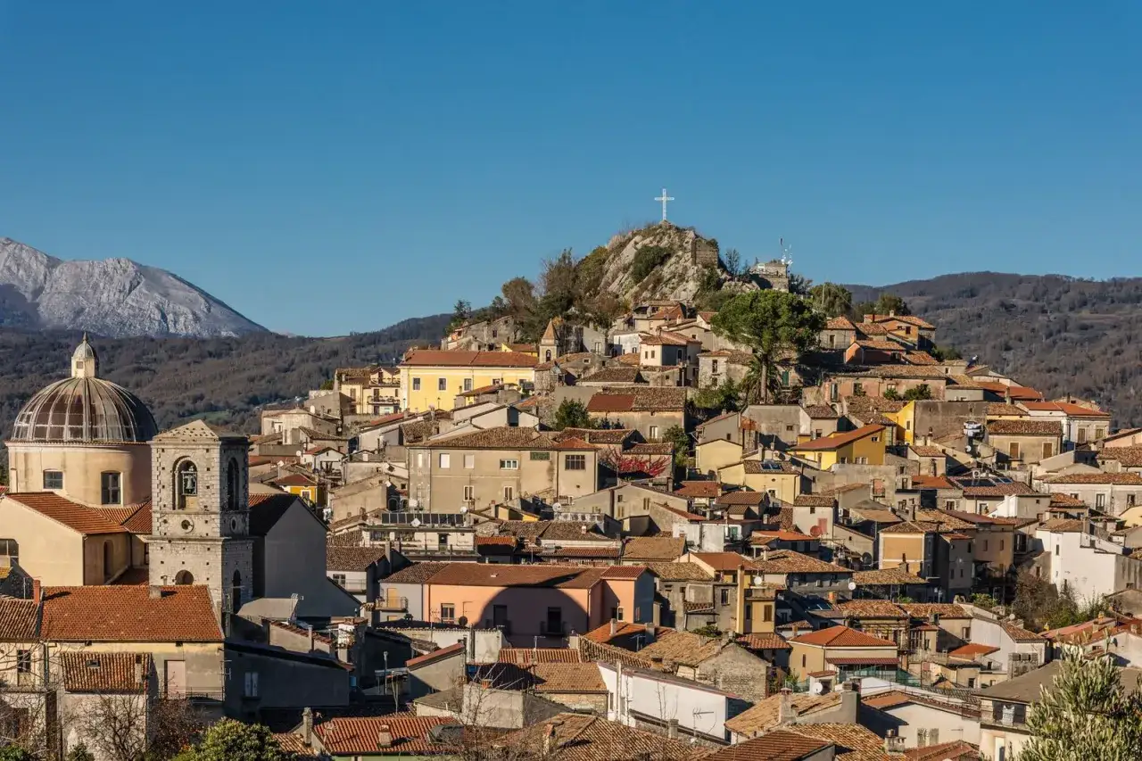 Borgo arroccato in Basilicata, con una chiesa a cupola e un campanile, dominato da una croce su una collina rocciosa.
