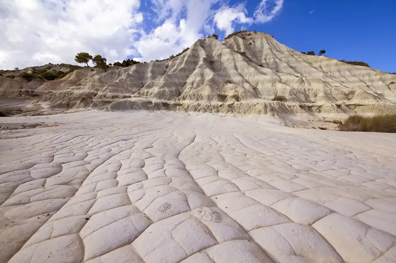 Paesaggio lunare con rocce erose e crepate, tipico delle colline senesi. Il terreno bianco e le forme scolpite dal vento creano un'atmosfera surreale sotto un cielo azzurro con nuvole bianche.