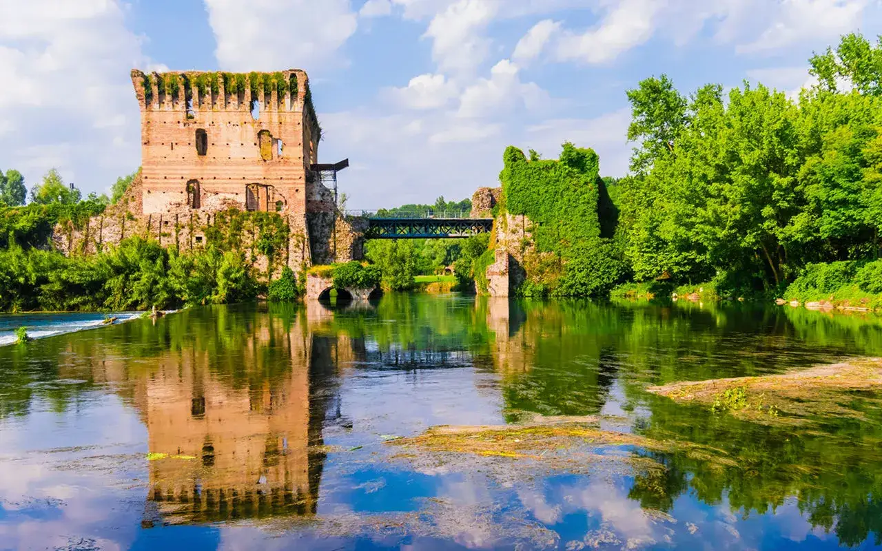 Rovine di un castello su un fiume in Italia, con un ponte metallico che collega le sponde. Un incantevole scorcio di una città sull'acqua.