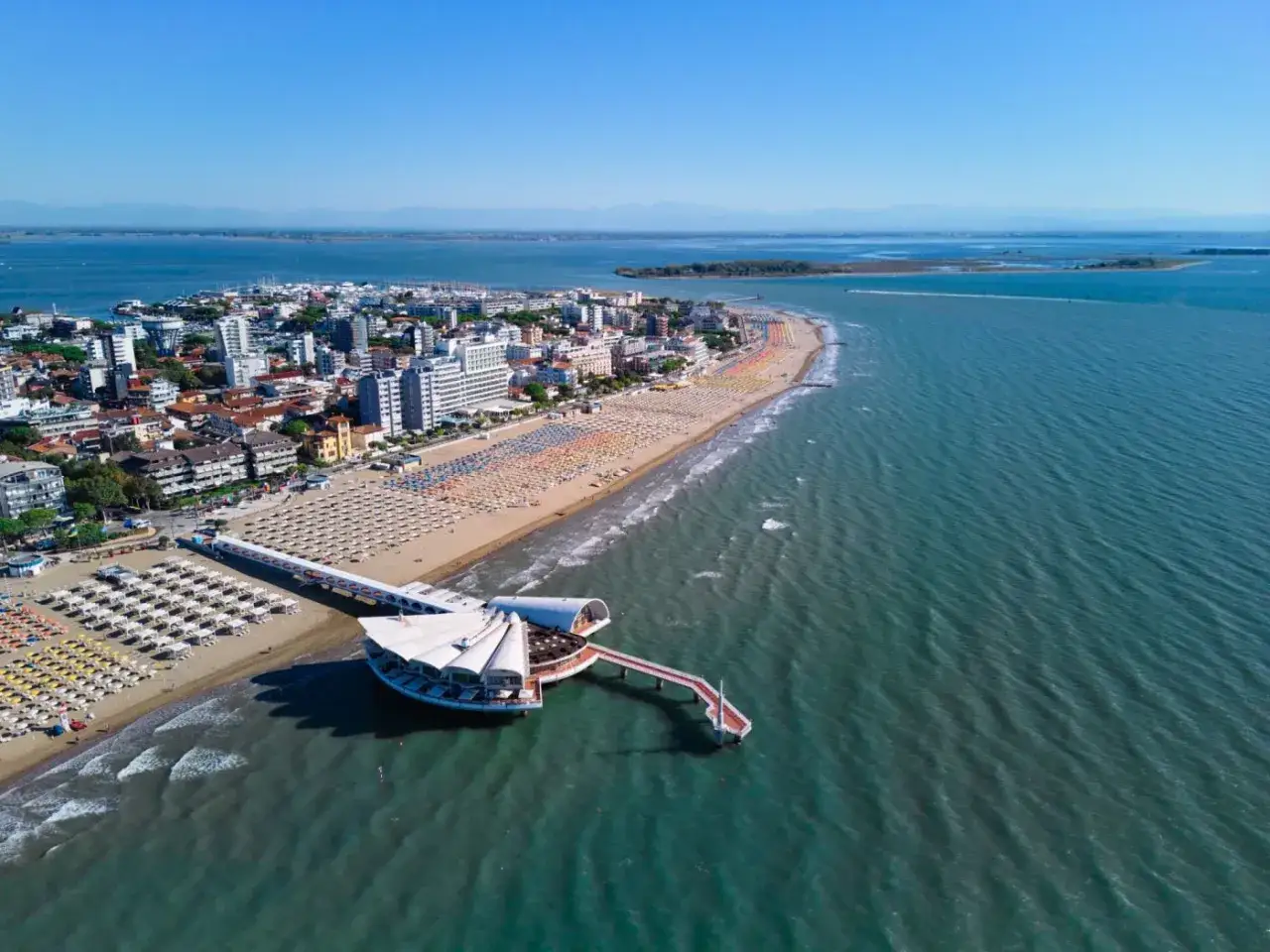 Un'ampia spiaggia attrezzata con ombrelloni colorati si estende lungo la costa del mare Friuli Venezia Giulia, con un moderno pontile che si protende sull'acqua.
