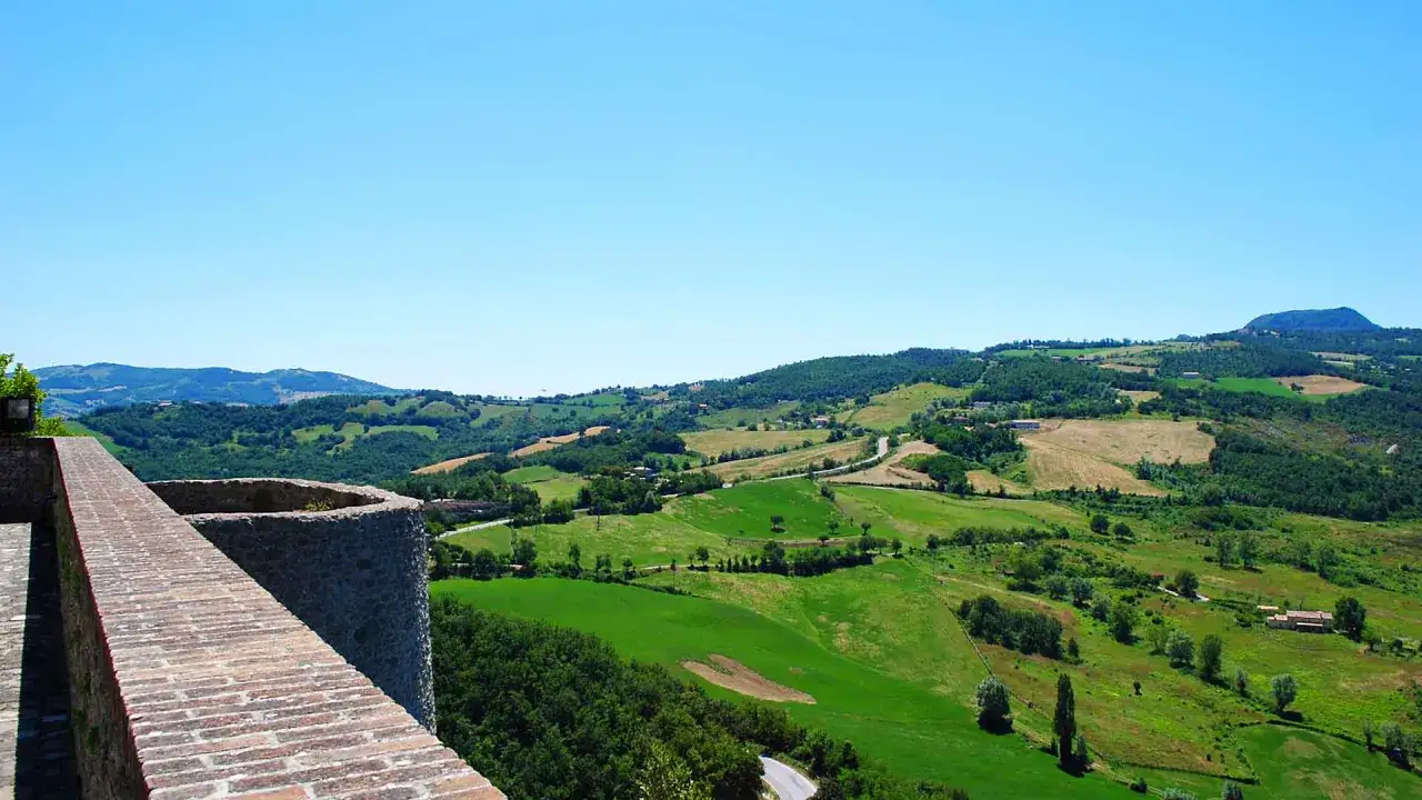 Vista panoramica collinare con campi verdi e alberi, con un muro di mattoni in primo piano. Il borgo San Leo si staglia sullo sfondo.
