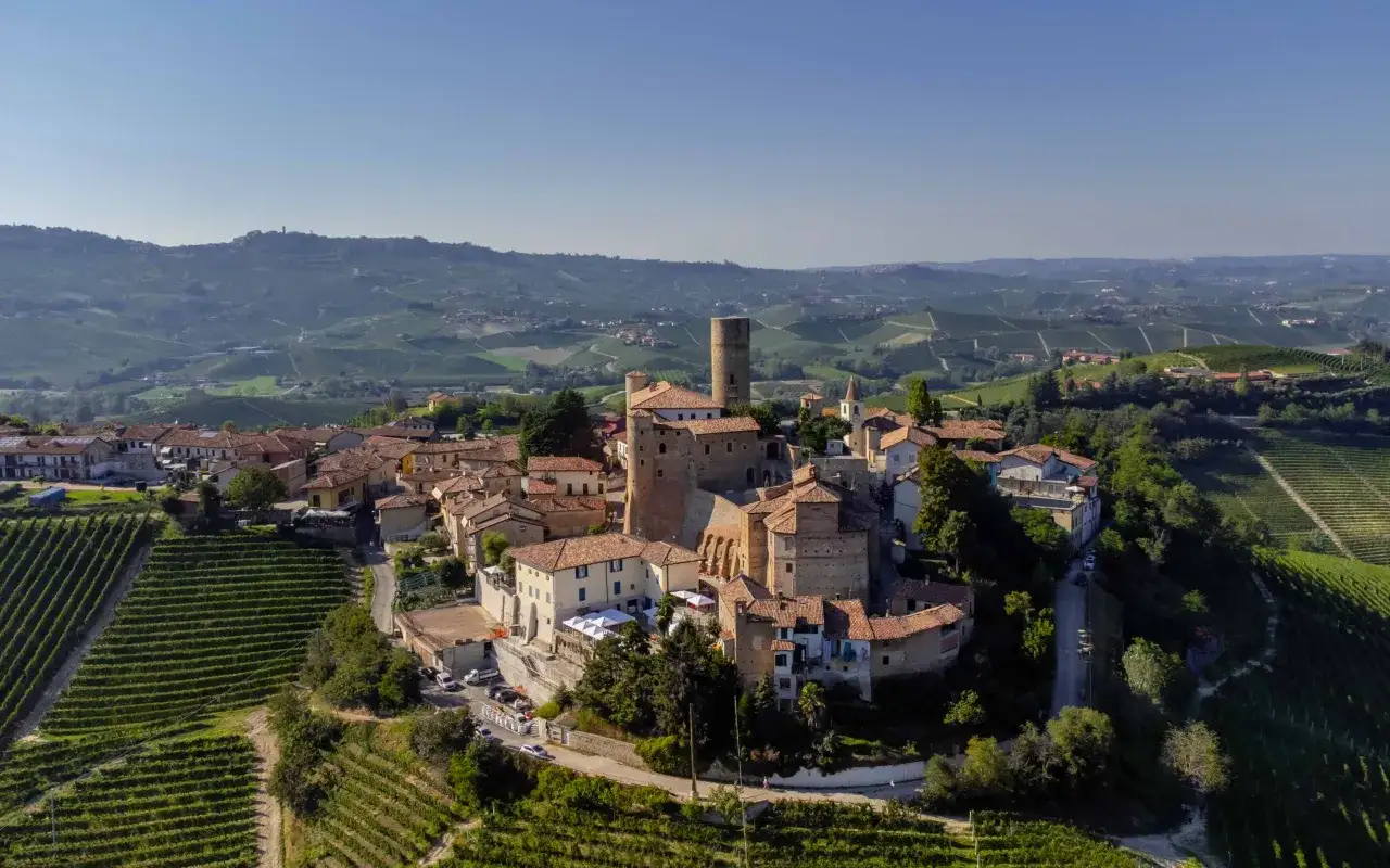 Borgo medievale tra le colline vitate dei paesi delle Langhe. Un castello domina il panorama, circondato da case con tetti rossi e vigneti a terrazze.