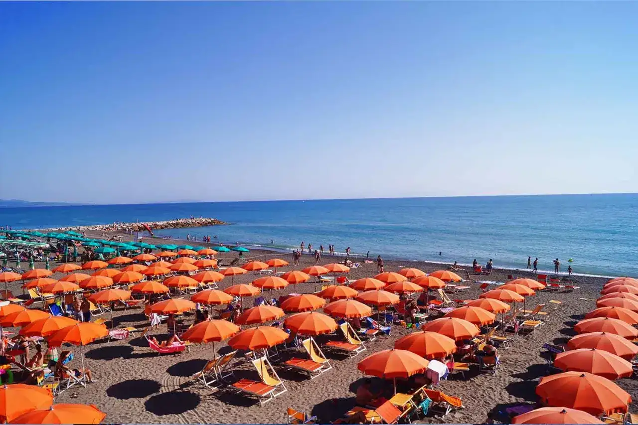 Spiaggia affollata con ombrelloni arancioni e verdi, lettini e persone che si godono il sole. Il mare azzurro e il cielo limpido creano una perfetta sassetta distanza dal mare.
