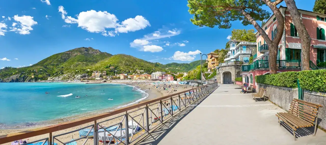 Splendida spiaggia sabbia Liguria con mare azzurro, colline verdi e case colorate.