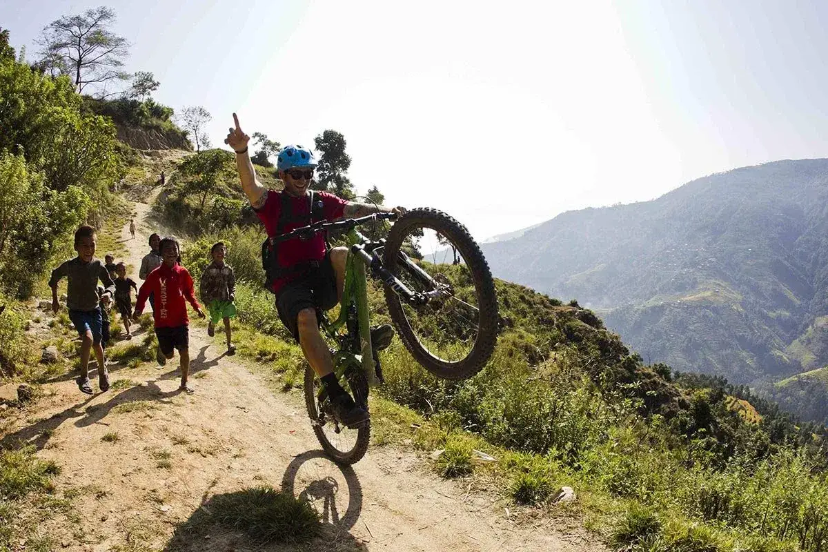 Uomo in mountain bike fa impennata, inseguito da bambini gioiosi su sentiero collinare. Guida all'acquisto perfetta per avventure così!