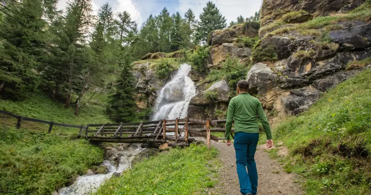 Escursionista ammira una cascata in montagna vicino Milano, estate. Sentiero e ponte in legno tra alberi e rocce.