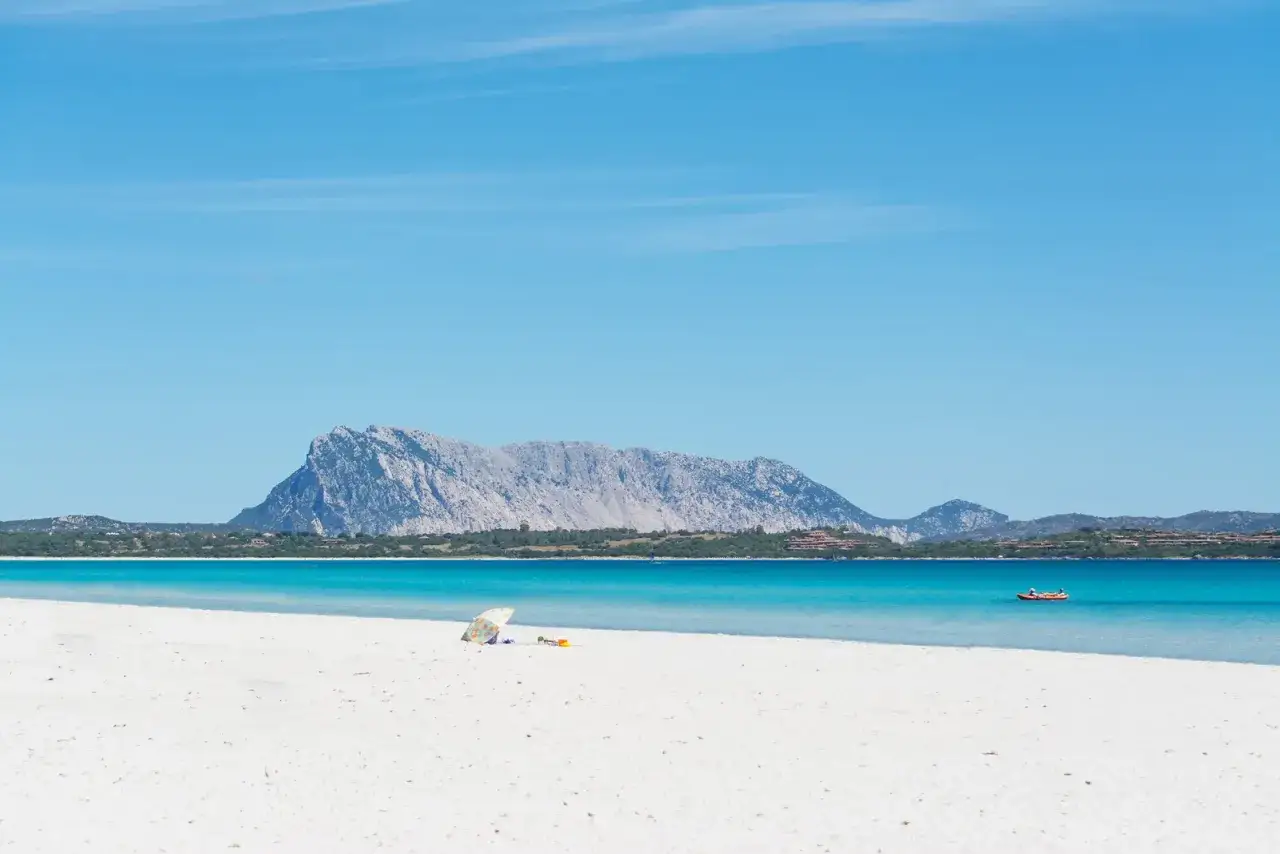 Spiagge di San Teodoro con sabbia bianca, mare cristallino e la sagoma imponente di Tavolara all'orizzonte. Un ombrellone colorato e un kayak invitano al relax.