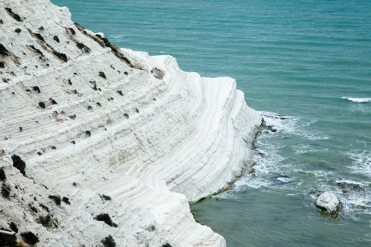 La scogliera bianca di Sicilia, scolpita dal mare, si tuffa in acque turchesi. Strati di roccia calcarea creano un paesaggio mozzafiato.
