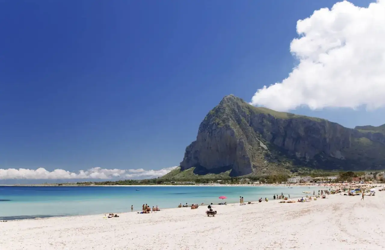 Spiaggia dorata e mare cristallino vicino Palermo, con una maestosa montagna che domina il paesaggio. Turisti godono del sole e delle acque limpide.