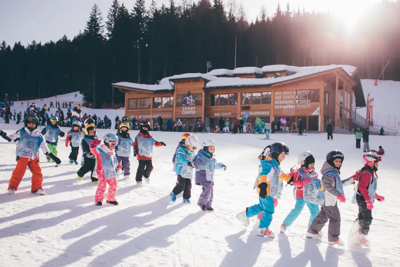 Bambini in fila, pronti dove imparare a sciare, davanti a un centro sportivo con scuola sci e noleggio.