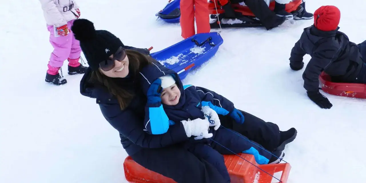 Mamma e bambino felici sciano in Abruzzo con slittino, circondati da neve e altri bambini su slitte.