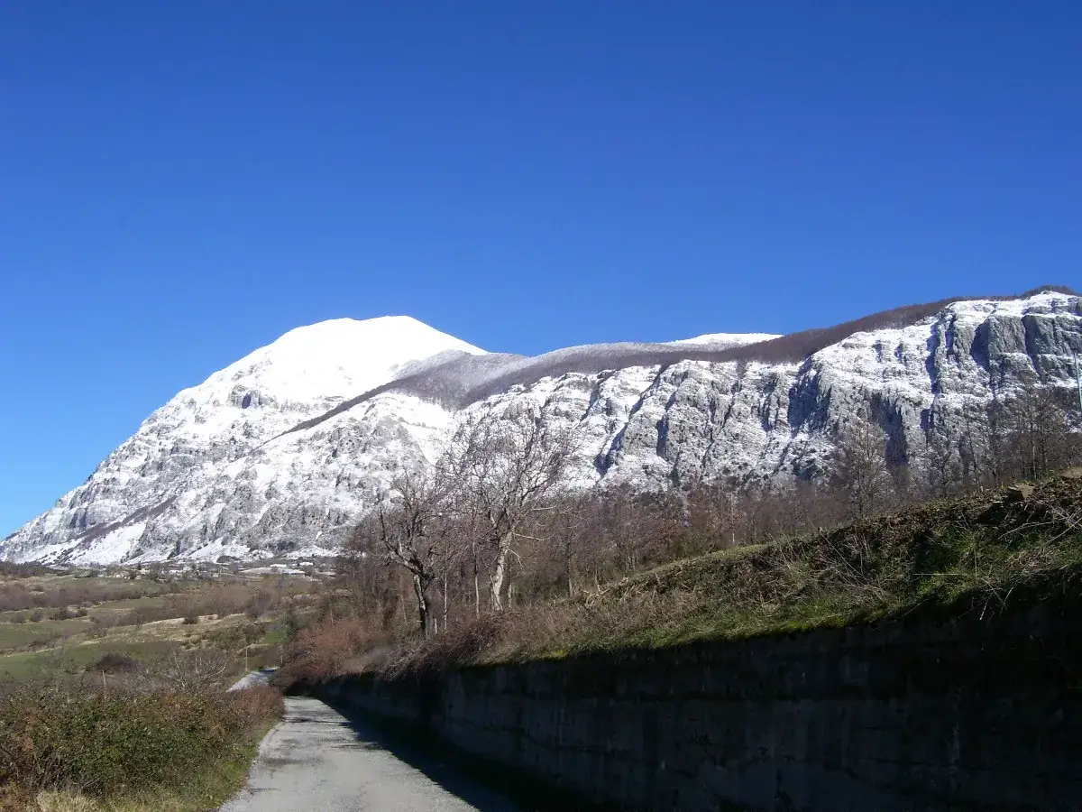 Strada sterrata con muro di contenimento e montagne innevate sullo sfondo, un paesaggio che mostra dove si trova Latronico.