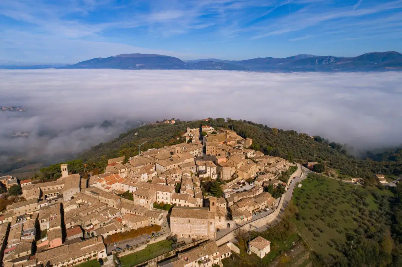 Un borgo medievale su una collina, avvolto dalla nebbia, con montagne sullo sfondo. Ecco cosa vedere a Montefalco, un gioiello umbro.