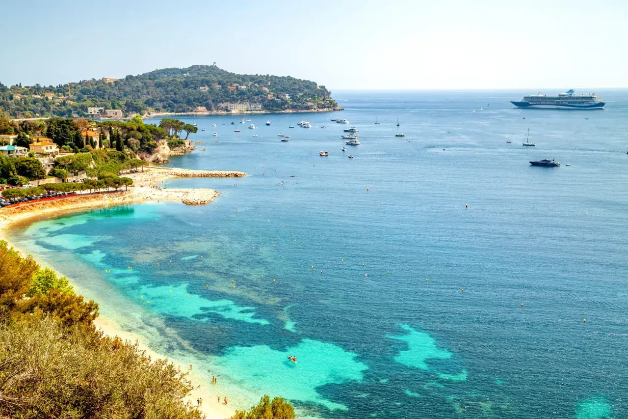 Vista panoramica della costa azzurra con mare cristallino, spiagge dorate, ville lussuose e yacht all'ancora.