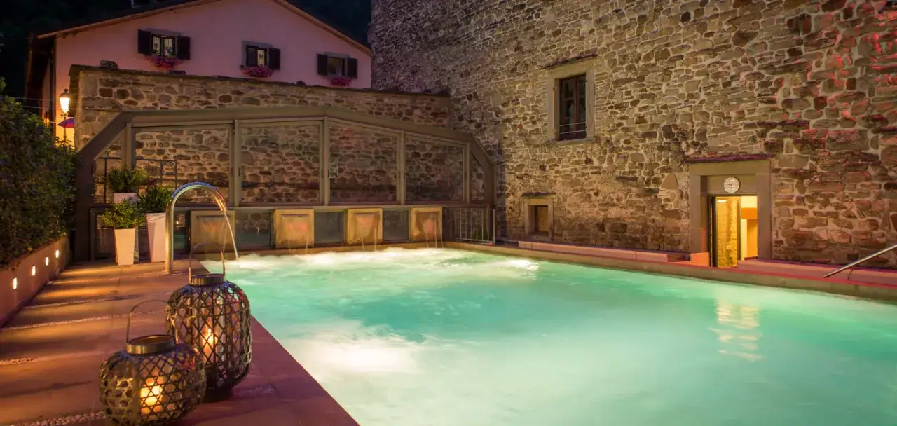 Piscina termale illuminata con cascate d'acqua, lanterne decorative e un edificio in pietra. Un'oasi di relax tra le migliori terme Emilia Romagna.