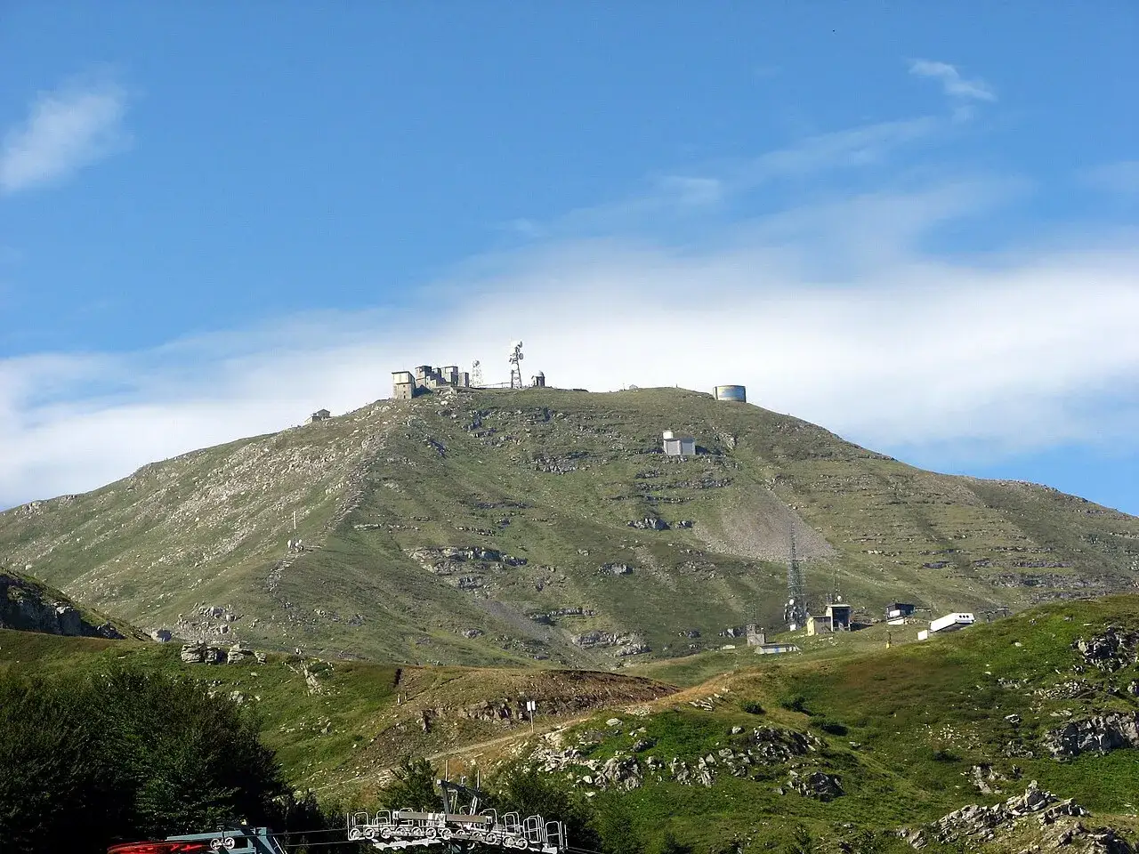 Il monte più alto dell'Emilia Romagna, con edifici e antenne sulla cima, sotto un cielo azzurro con nuvole bianche.