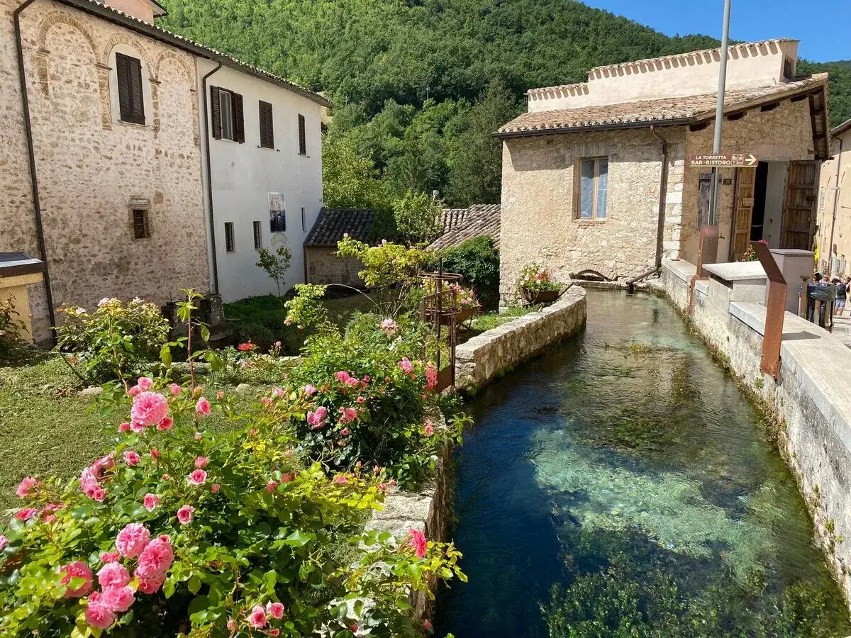 Un ruscello attraversa un borgo vicino Nocera Umbra, con case in pietra e rose rosa in fiore.