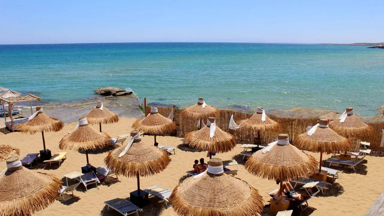 Ombrelloni di paglia e lettini sulla sabbia dorata, con il noto mare azzurro che si estende all'orizzonte.