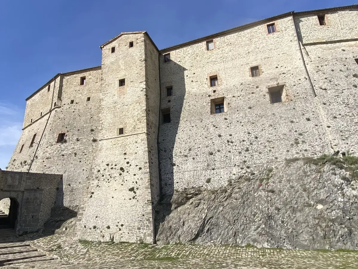 Imponente rocca di Borgo San Leo su uno sperone roccioso, con mura in pietra chiara e piccole finestre quadrate sotto un cielo azzurro.