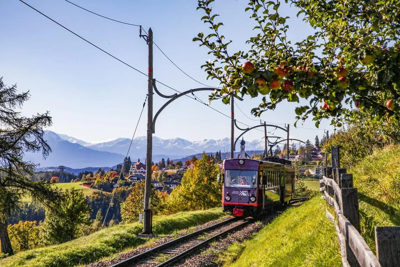 Il trenino del Renon attraversa un paesaggio autunnale con montagne innevate sullo sfondo. Un ramo di melo carico di frutti fa da cornice.
