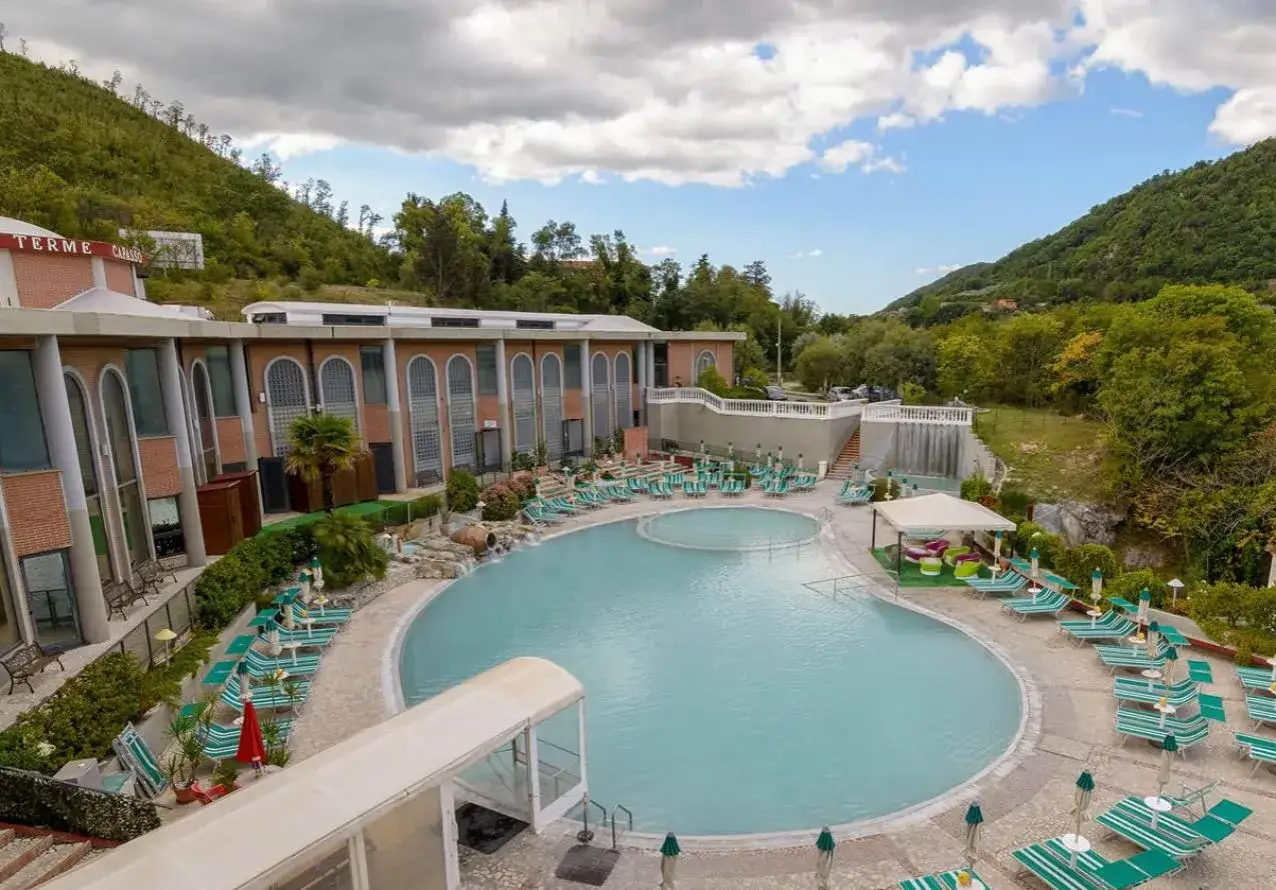 Piscina termale con lettini e ombrelloni, circondata da un edificio in mattoni e colline verdi. Un'oasi di relax, perfetta per godersi le contursi terme gratis.