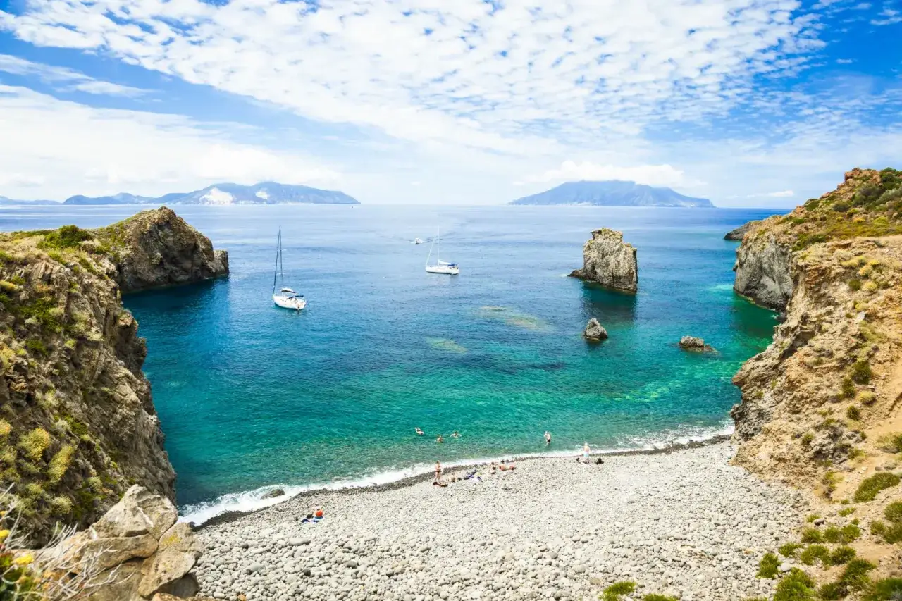 Spiagge di ciottoli e acque cristalline alle Isole Eolie, con barche a vela e persone che nuotano.