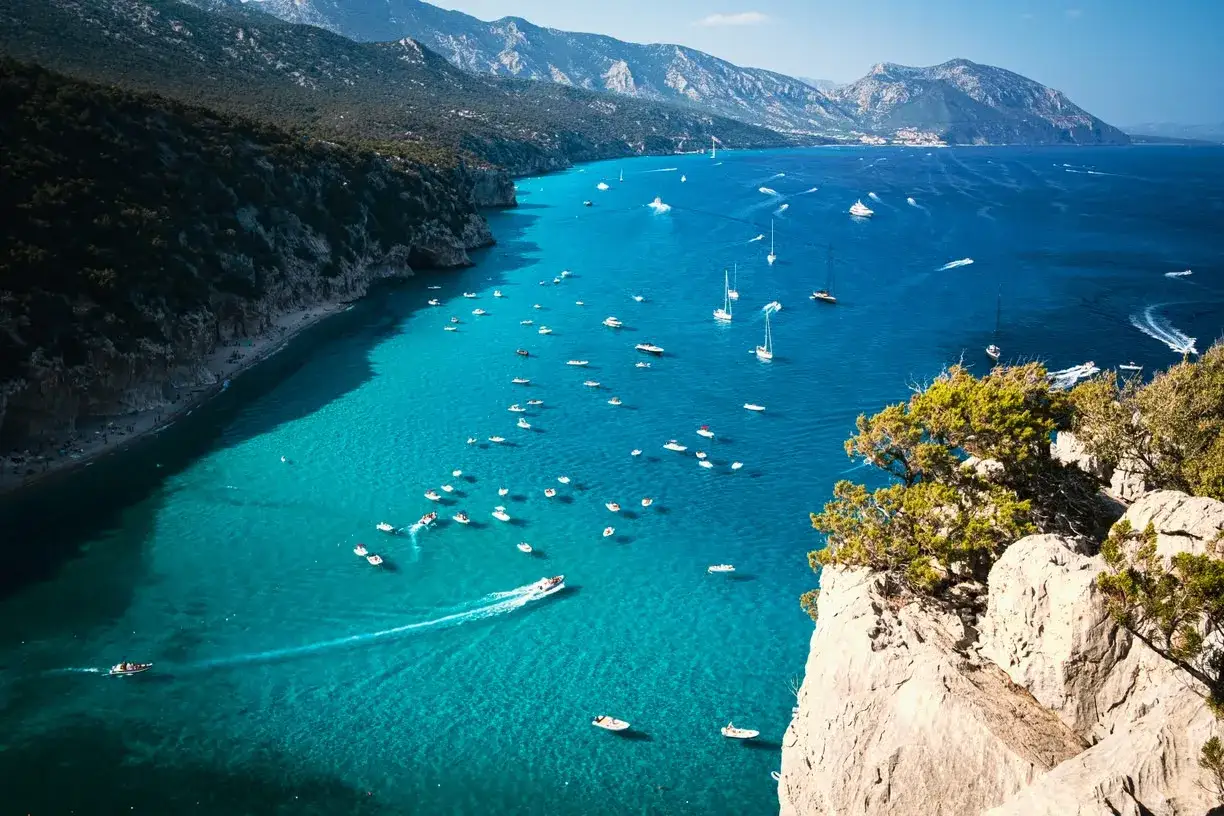 Un mare cristallino e tante barche animate le coste della Sardegna, con montagne verdi a fare da sfondo.