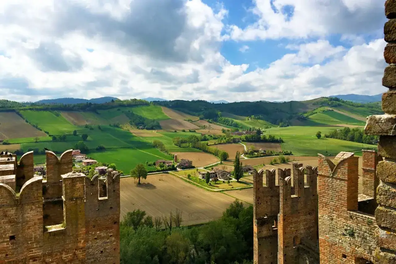 Torri di un borgo medievale a Piacenza, con dolci colline verdi e campi coltivati sotto un cielo nuvoloso.