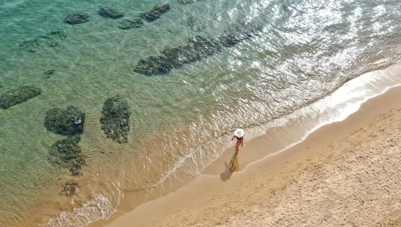 Una donna con cappello bianco cammina sulla sabbia dorata, bagnata dalle onde cristalline del mare. Un angolo di paradiso vicino a Roma.