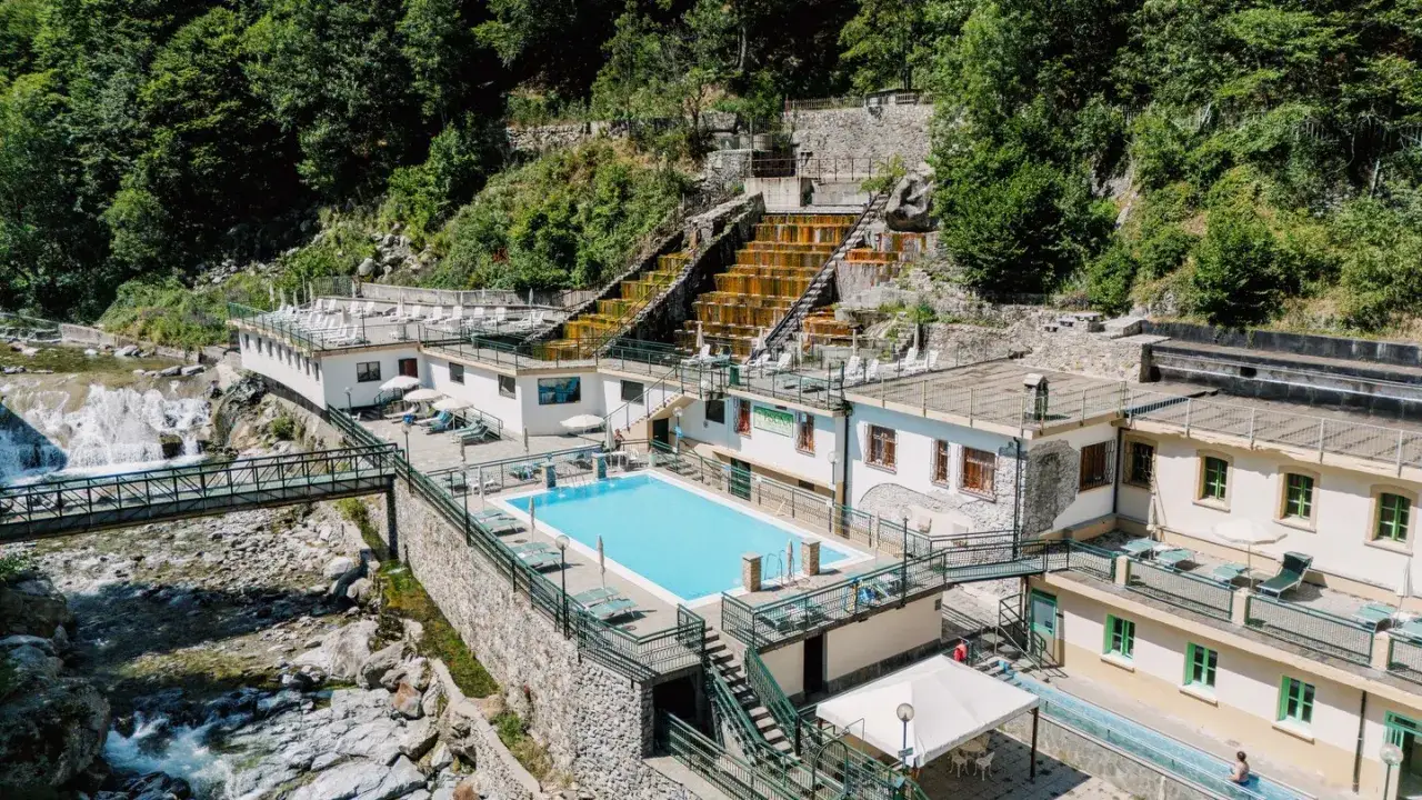 Piscina e cascate naturali alle terme di Valdieri, con edifici bianchi e vegetazione lussureggiante.