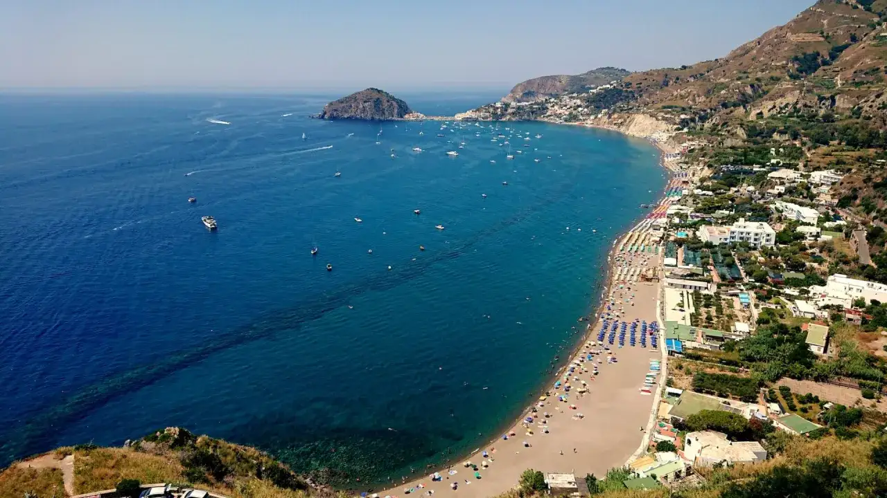 Vista aerea di una baia con spiaggia affollata, ombrelloni colorati e mare blu intenso. Un promontorio roccioso e un isolotto punteggiano l'orizzonte, evocando le 5 spiagge più belle di Ischia.