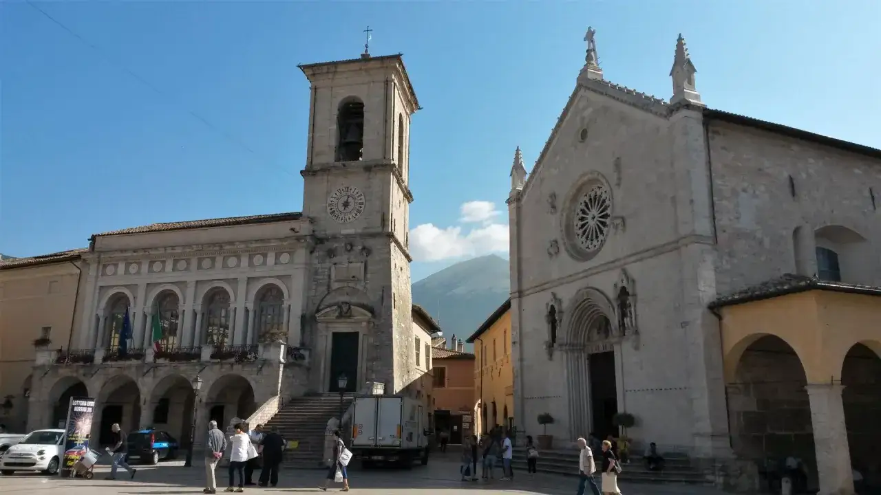 Piazza di Norcia, famosa per la sua bellezza e storia, con il campanile e la chiesa che si stagliano sotto un cielo azzurro.