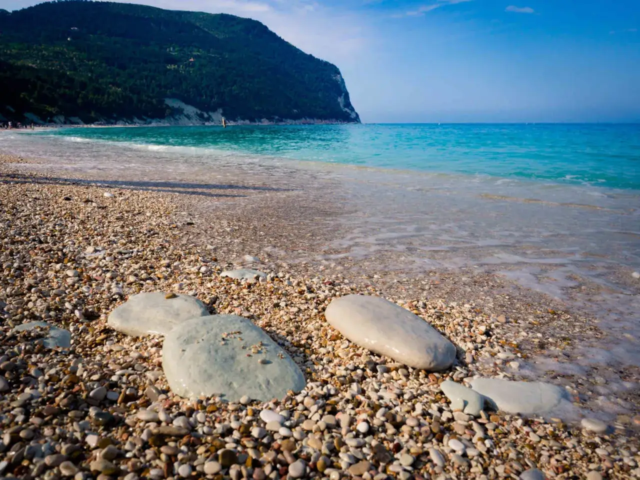 Spiaggia di sassi con mare turchese e il Monte Conero dove si trova.