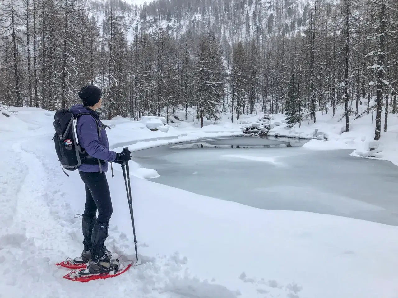 Donna con ciaspole ammira un lago ghiacciato durante le ciaspolate in Val d'Aosta, circondata da un bosco innevato.