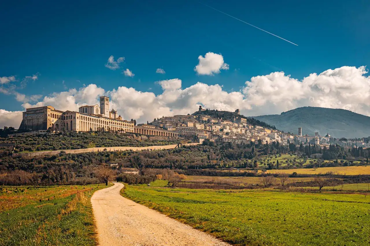 Un sentiero sterrato conduce verso la collina dove si trova Assisi, con la sua imponente basilica e il borgo medievale sotto un cielo azzurro.