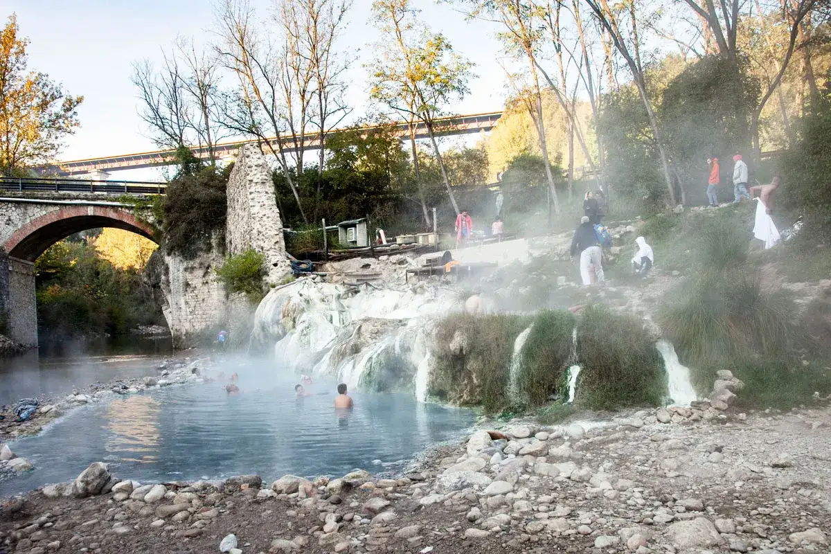 Persone si rilassano nelle acque fumanti delle terme di Petriolo, con un antico ponte sullo sfondo.