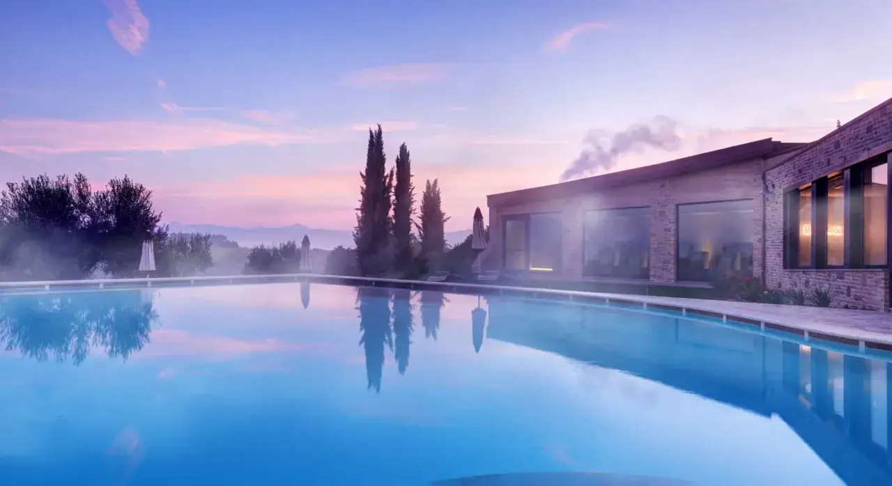 Piscina a sfioro con riflessi del cielo rosa e viola, cipressi e un edificio moderno. Un'oasi di relax nelle terme di Perugia.