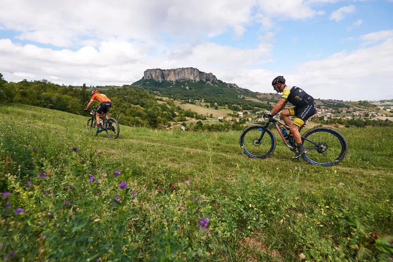 Cicloturismo in Emilia Romagna: due ciclisti affrontano un sentiero erboso con un suggestivo borgo sullo sfondo.