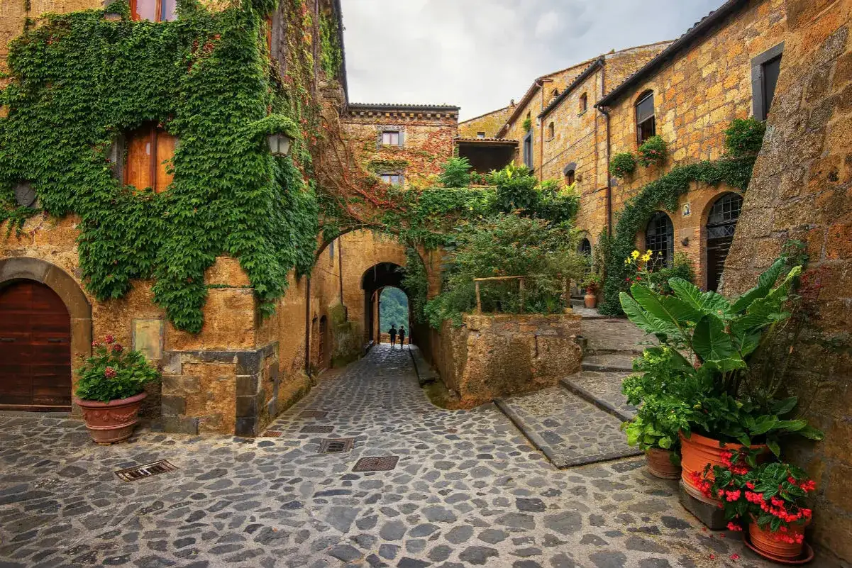 Antichi scorci di borghi medievali, con case in pietra ricoperte d'edera e un vicolo acciottolato che conduce a un arco.