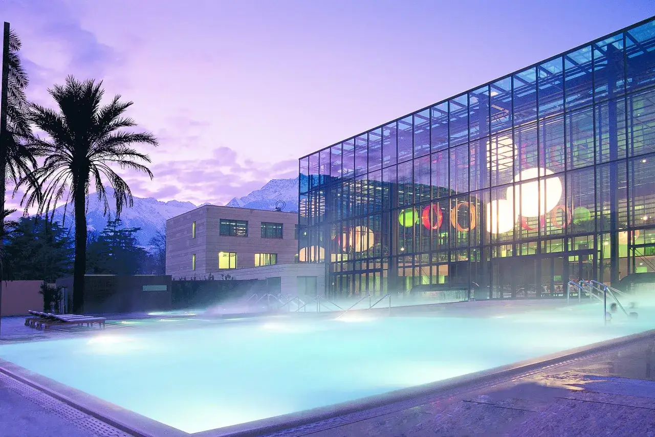 Piscina esterna illuminata con vapore acqueo alle Merano Terme, con palme e montagne innevate sullo sfondo al tramonto.