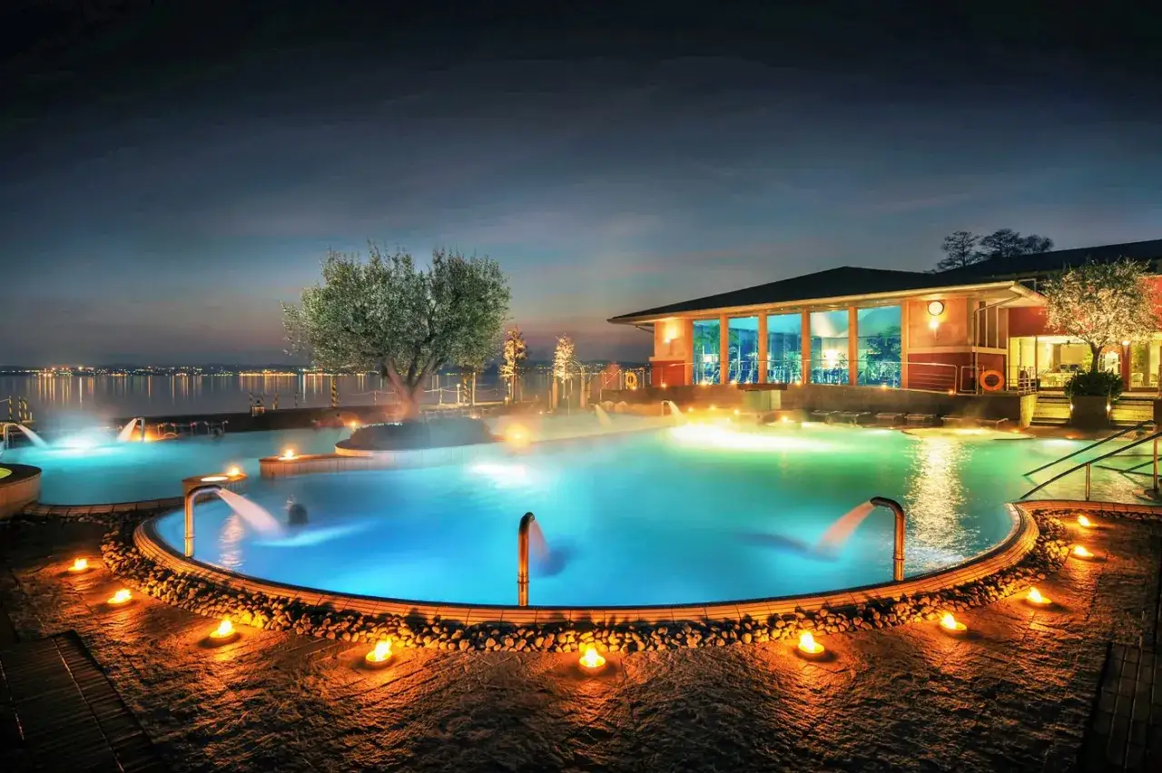 Piscine termali illuminate da candele e luci soffuse, con un albero fiorito e un edificio moderno sullo sfondo. Un'oasi di relax che ricorda le terme a Brescia.